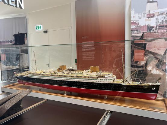 Ein detailliertes Modell des Ozeandampfers "Bremen" in einer Vitrine, begleitet von Hintergrundbildern und Informationstafeln über das Schiff. Das Modell verfügt über einen schwarz-roten Rumpf mit goldenen Akzenten.