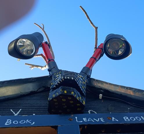 book box gargoyle, a horned rabbit creature, with solar lights on the end of each horn, pointed back down at interior of box.

Incomplete view of phrases painted in white on box door, 

"Take a book, leave a book"