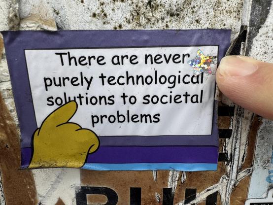 A sticker that says ‘there are never purely technological solutions to societal problems”.  The art on the sticker is a scene from The Simpsons when a bus driver points at a sign in the front of the bus.
