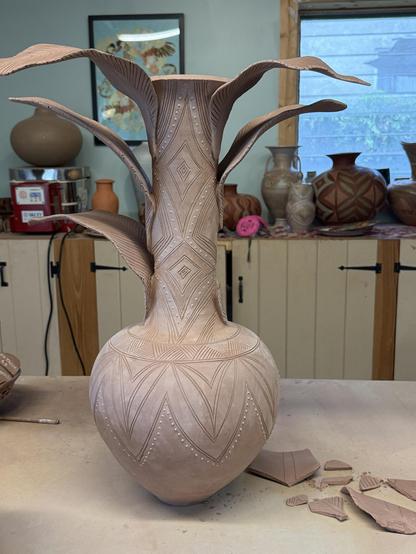 A large unfired vase with a very tall neck, with sculpture leaves attached to it—three on each side. The neck and body of the vase feature carved geometric decorations. The entire piece is bone dry—a pale ashy tone. The lower leaf on the right side has fallen and is in pieces beside it on my work table. A small kiln, other finished pots and a window can be seen in the background.