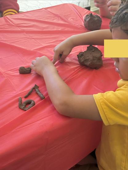 A child is engaged in sculpting with brown clay on a red plastic tablecloth. The child is shaping the clay into various forms, including the numbers 6 and 7. Other clay pieces are also present on the table.