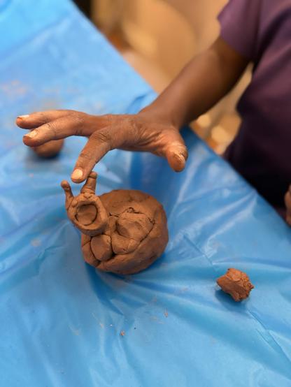 A hand is shaping a small snail out of brown clay on a blue table surface, with additional clay pieces nearby.