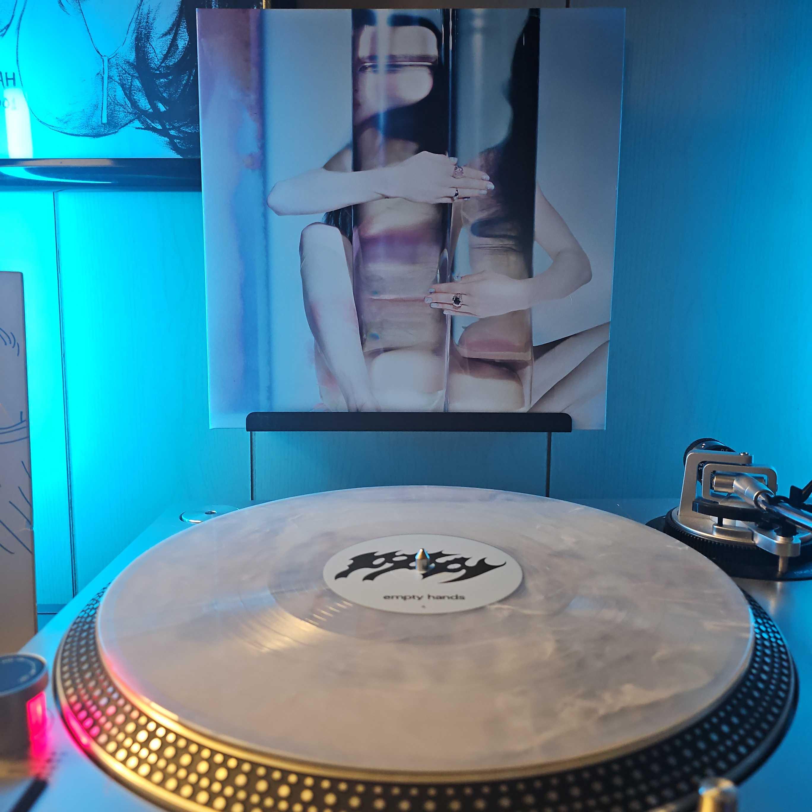 A White [Iridescent] With Blue Marble vinyl record sits on a turntable. Behind the turntable, a vinyl album outer sleeve is displayed. The front cover shows Poppy sitting behind a large glass cylinder, which she is wrapping her arms around. Her face and body are distorted through the glass