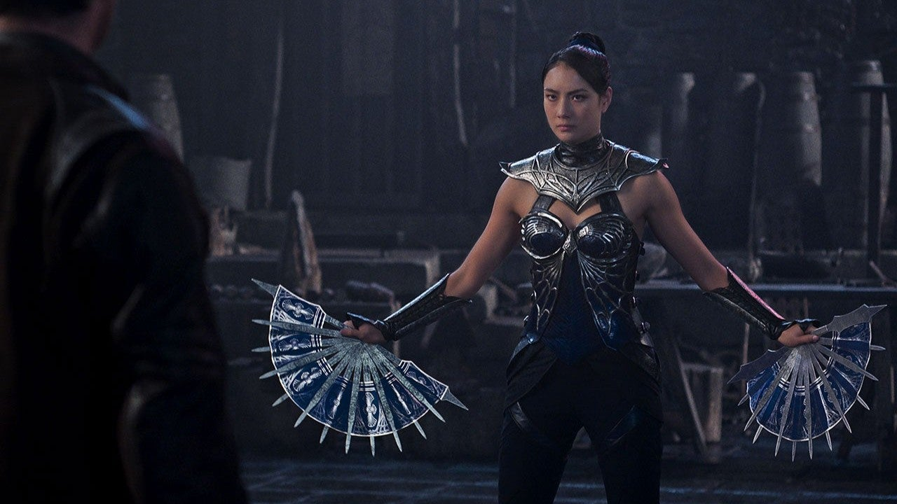 Screenshot of scene from Mortal  Kombat II (2026) movie showing Princess Kitana (Asian woman wearing blue melal body armor on her shoulders and neck, going down to a metal bikini top and blue pants and carrying blue metal battle fans)