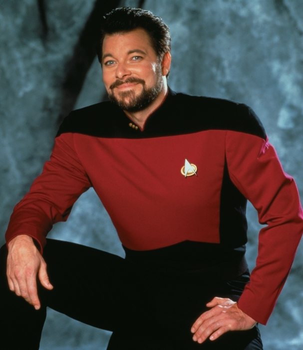 promo photo of William Riker (bearded white man in his 30s in the red and black Star Trek The Next Generation command uniform) sitting with one leg up and his hand resting on his knee and the other on high thigh