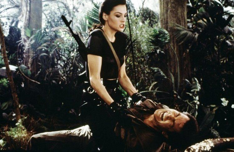 photo from Goldeney of Xenia Onatopp (white woman with dark hair in a black t-shirt and black pants with a gun on her back) straddling James Bond (Pierce Brosnan) in a jungle as he screams in pain
